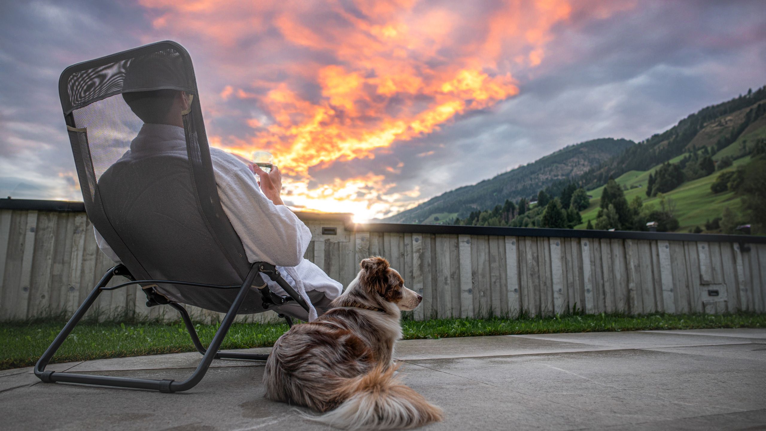 Gast im Apartmenthotel Dolomit Royal in Osttirol entspannt auf der Terrasse mit Hund bei Sonnenuntergang – Blick auf die Berge und malerische Alpenlandschaft.
